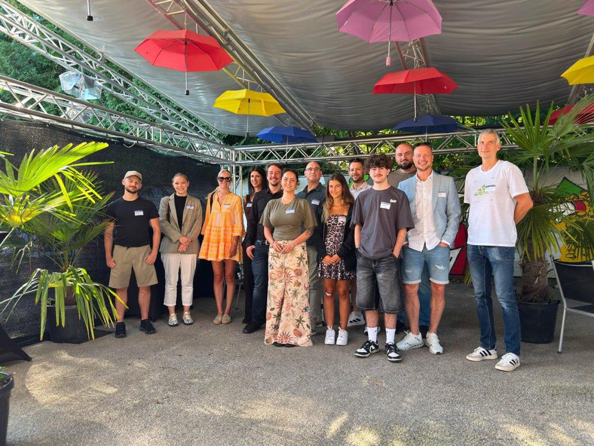 Vierzehn Teilnehmerinnen und Teilnehmer eines KINN-Treffs stehen im Freien unter bunten aufgespannten Schirmen für ein Gruppenfoto