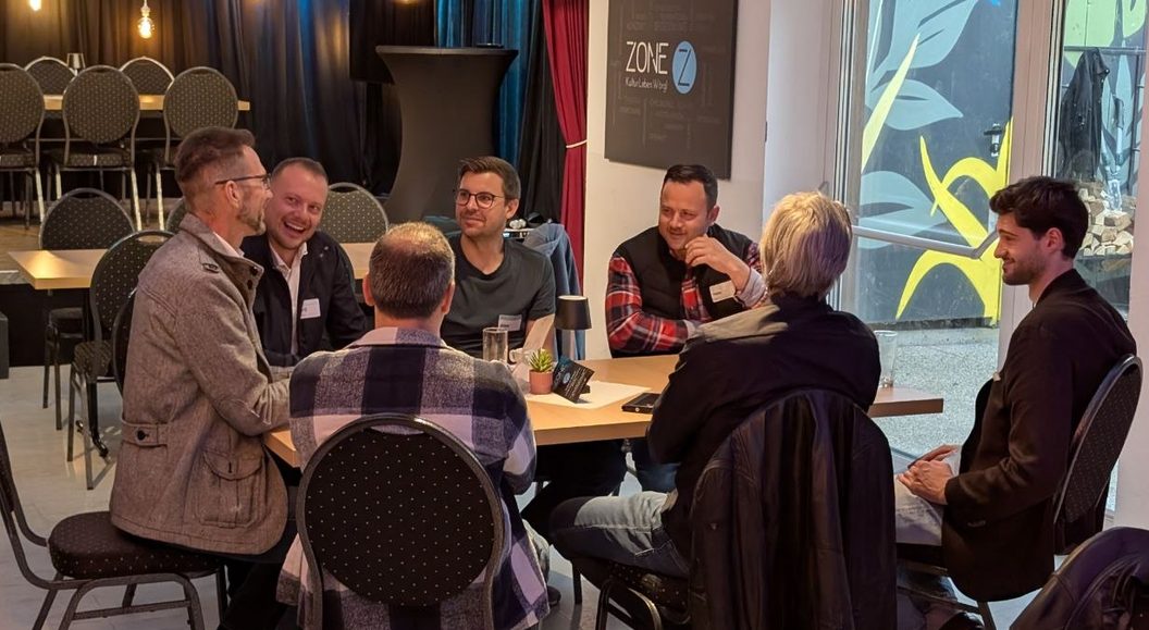 Foto: Sieben Menschen sitzen um einen Tisch in der ZONE in Wörgl, reden und lachen miteinander, vor ihnen Gläser und eine kleine Pflanze. Einige tragen Namensschilder. An der Wand dahinter ein dunkles Schild mit der Aufschrift ZONE, Kultur Leben Wörgl. Links leere gepolsterte Stühle an langen Tafeln, rechts eine Glastür mit blauen und gelben Pflanzenmotiven.